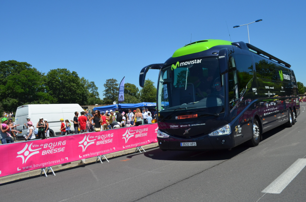 Ein schwarzer und grüner Bus fährt auf einer Straße mit einer Menschenmenge, einige tragen Mützen, ein Banner mit Text auf der linken Seite, Bäume und einen klaren blauen Himmel im Hintergrund.