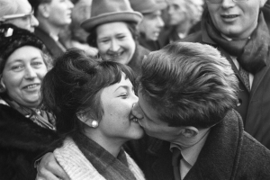 Ein Schwarz-Weiß-Foto von einem Mann und einer Frau, die leidenschaftlich küssen, während sie von einer lachenden Menge umgeben sind.