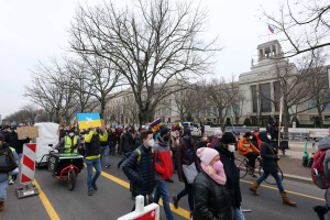 Eine große Gruppe von Menschen nimmt an einer Protestdemo auf einer Straße in Washington, D.C., teil, einige halten Schilder und Transparente, andere fahren Fahrräder, und Schilder, Bäume und ein klarer blauer Himmel sind im Hintergrund zu sehen.
