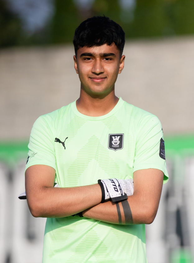 Ein junger Mann in einem grünen Hemd steht auf einem Fußballfeld und schaut mit verschränkten Armen und einem Lächeln in die Kamera, während der Hintergrund unscharf ist.