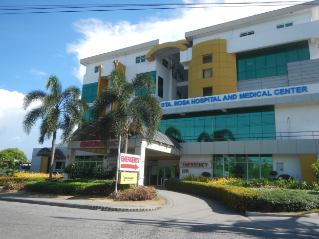 Außenansicht des St. Rosa-Krankenhauses und -Medizinischen Zentrums, ein modernes Gebäude mit Glasfenstern und Beschriftung, umgeben von Bäumen und Gras mit einer Straße im Vordergrund und einem bewölkten Himmel darüber.