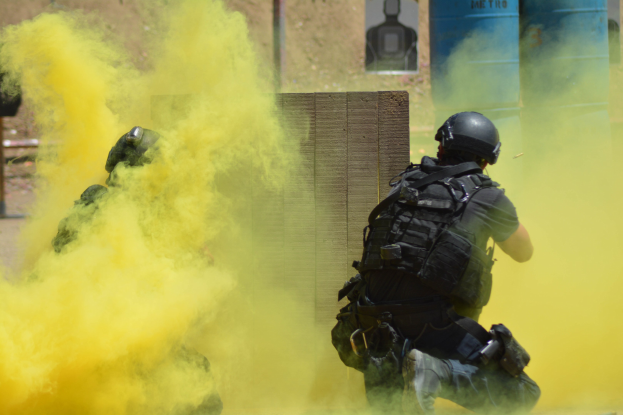 Zwei Polizisten in Einsatzausrüstung mit gelbem Rauch aus dem Mund, die an einer Wand mit Texttafel und anderen Gegenständen im Hintergrund stehen.