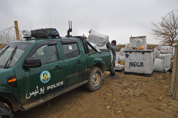 Polizeilaster auf einem Schotterweg neben einem Maschendrahtzaun geparkt, mit Text und Logo auf der Seite, zwei Personen in der Nähe und Hintergrundelemente wie Taschen, Baum, Pfahl und Himmel.