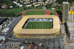 Luftaufnahme eines Fußballstadions im Zentrum einer Stadt, umgeben von Gebäuden, Bäumen, Straßen, Fahrzeugen, Masten und Kränen, mit saftig grünem Rasen.