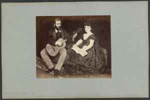 Ein Mann im Anzug und eine Frau, beide halten Gitarren und Papierstücke in den Händen und sitzen friedlich nebeneinander.