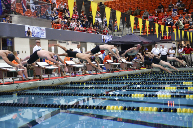 Schwimmer tauchen in ein Schwimmbecken, während Zuschauer von Stühlen und Tischen aus zuschauen, mit Bannern, Fahnen und einem Zelt im Hintergrund.