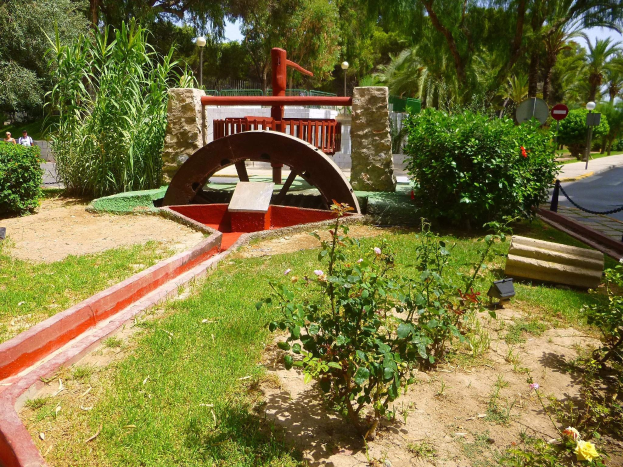 Minigolfanlage in einem Park mit Grünflächen, einer Straße mit Fußgängern und Schildern sowie Bäumen vor einem blauen Himmel.