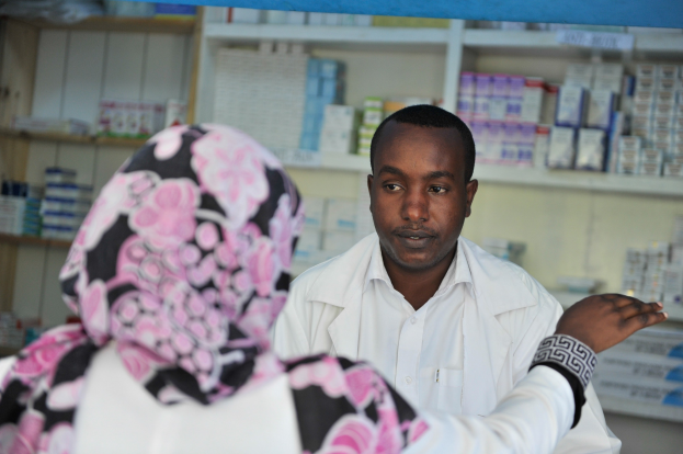 Ein Apotheker in einem weißen Laborkittel steht einer sitzenden Frau in einer Apotheke gegenüber, mit Regalen voller Schachteln und Gegenständen im Hintergrund.