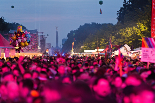 Eine große Menge bei einem nächtlichen Musikfestival mit bunten Lichtern, Ballons, Zelten, Schildern, Fahnen, Bäumen, einem Turm und einem sternenklaren Himmel im Hintergrund.