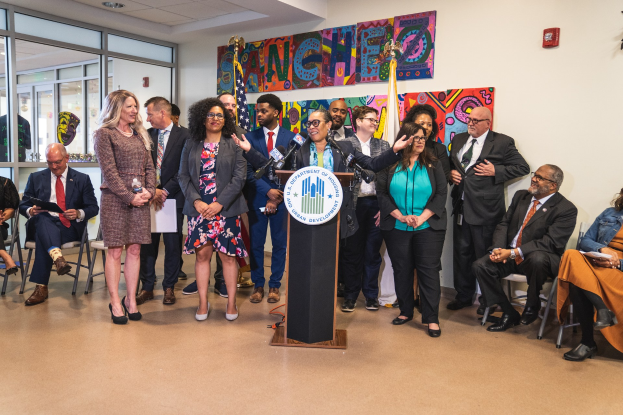 Gruppe von Menschen vor einem Rednerpult mit Mikrofon und einer Tafel mit Text, einige sitzen auf Stühlen und andere stehen, mit Flaggen, einem Feuerlöscher und Deckenleuchten an der Wand im Hintergrund.