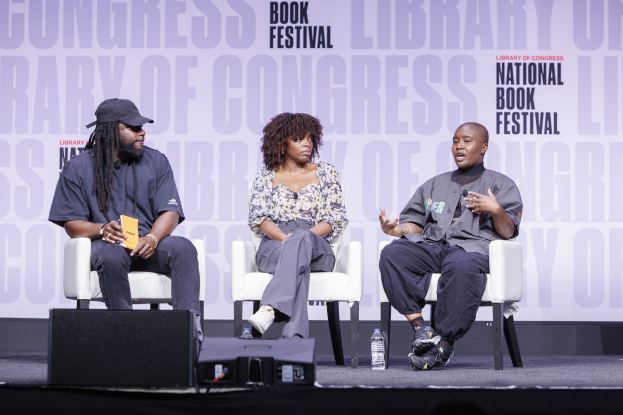 Drei Personen sitzen auf der Bühne der Library of Congress Book Festival, wobei ein Mann mit einer Mütze ein Buch hält und eine Frau in die Mitte spricht; Flaschen und andere Gegenstände sind auf der Bühne sichtbar.