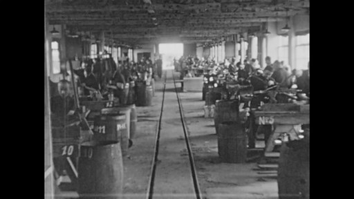 Schwarzes und weißes Foto einer Fabrik mit Arbeitern, die Maschinen bedienen, nummerierten Fässern, einer Eisenbahnschiene, Säulen und Deckenlampen während der industriellen Revolution.