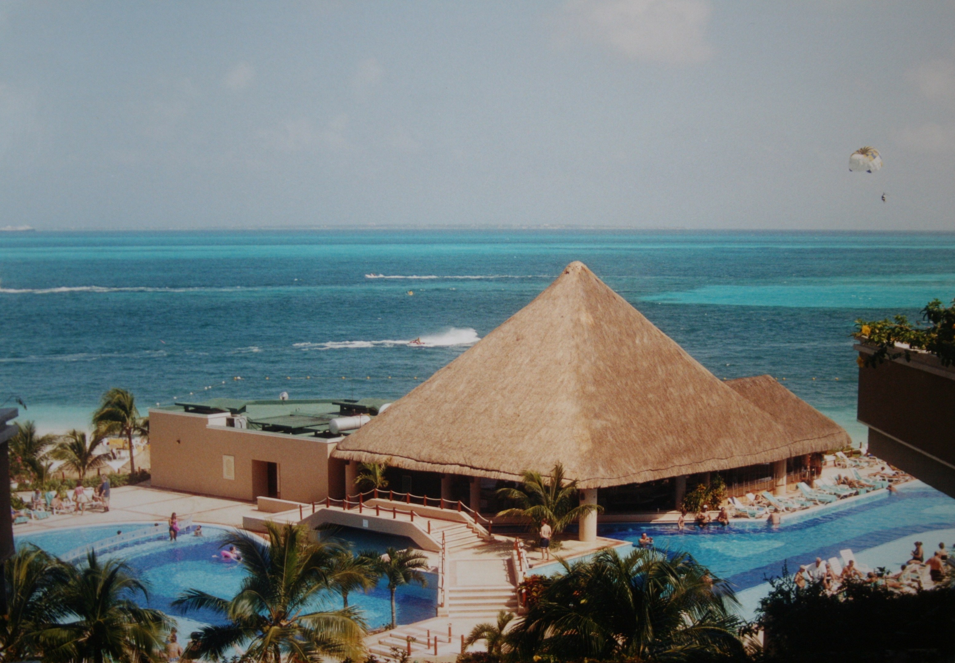 Resort mit einem gro├čen Schwimmbad, umgeben von Palmen, Hütten und Gel├Ąndern, mit Menschen im Pool, einem Ozean im Hintergrund, einem bew├Âlktem Himmel und einem Fallschirm über dem Pool.