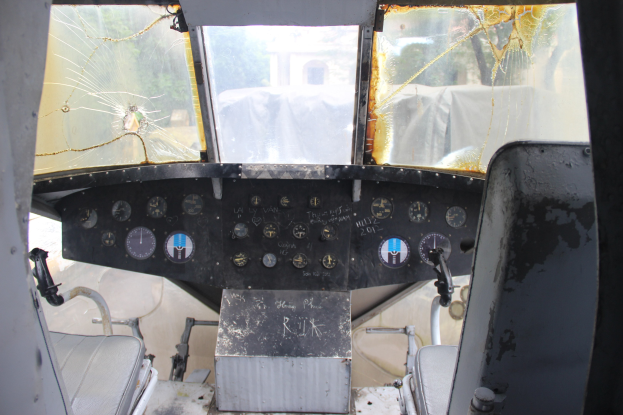 Cockpit-Innenseite eines Flugzeugs mit zerbrochener Frontscheibe, zwei Pilotensitzen und mehreren Kontrollinstrumenten, die durch das zerbrochene Glas hindurch Bäume und ein Gebäude zeigen.