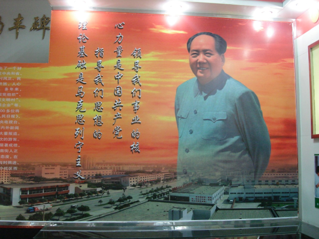 Großes Mao-Zedong-Plakat an einer Gebäudewand, beleuchtet von Lichtern, mit einem Foto-Rahmen und Text rechts daneben.