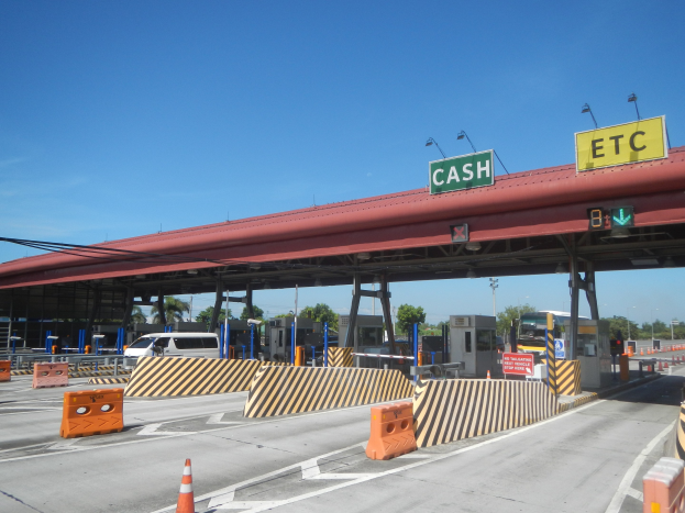 Mautstelle mit einem "Bar" Schild, Fahrzeugen, Sperrpoller, Schilder, Straßenlaternen, Strommasten, Stromkabel, Bäumen und einem bewölkten Himmel im Hintergrund.