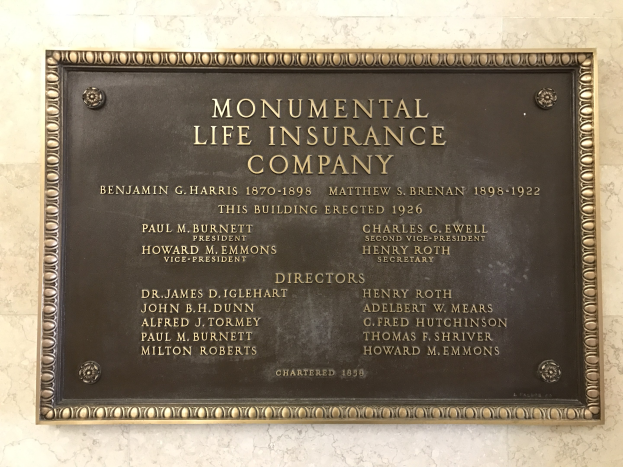 Plakette an einer Gebäudewand mit der Aufschrift "Monumental Lebensversicherungsgesellschaft" in fetter Schrift mit einem dekorativen Rahmen, auf dunklem Material montiert.