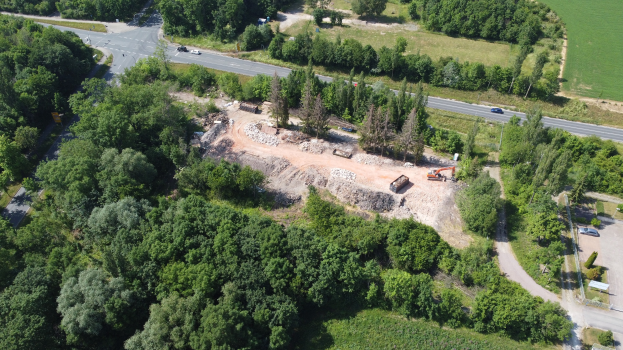 Luftbild einer Baustelle in einem bewaldeten Gebiet, das Bäume, Fahrzeuge auf einer Straße und Grasland zeigt.