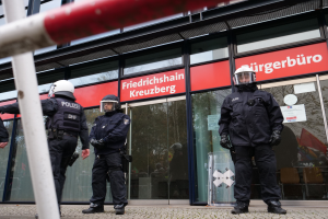 Eine Gruppe von Polizisten in Helmen und Handschuhen steht vor einem Glasgebäude, mit einem Pfahl links und Schildern im Hintergrund, die "Friedrichshain Kreuzberg" und Papiere an den Glaswänden zeigen.