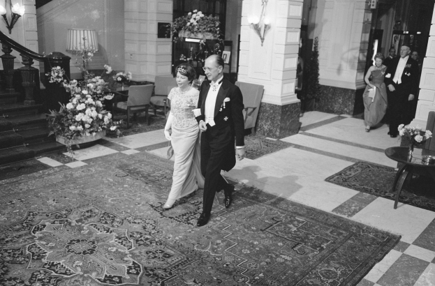 Schwarz-weiß-Foto einer Braut und eines Bräutigams, die durch die Lobby eines Hotels gehen, umgeben von Gästen, mit einem Blumenstrauß auf einem Tisch, Stühlen, Topfpflanzen, Lampen, einer Treppe und einem Geländer.