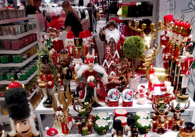 Ladeninnere voller Weihnachtsdekorationen, darunter Spielzeug, Tassen und andere Gegenst├Ąnde auf Tischen und Regalen, mit Menschen, Lichtern und einer Wand im Hintergrund.