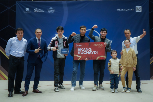 Gruppe von Menschen auf einer Bühne mit einer Tafel, auf der "Akang Kuchaydi - Zakovat Quiz" steht, im Hintergrund ein Banner und Lichter