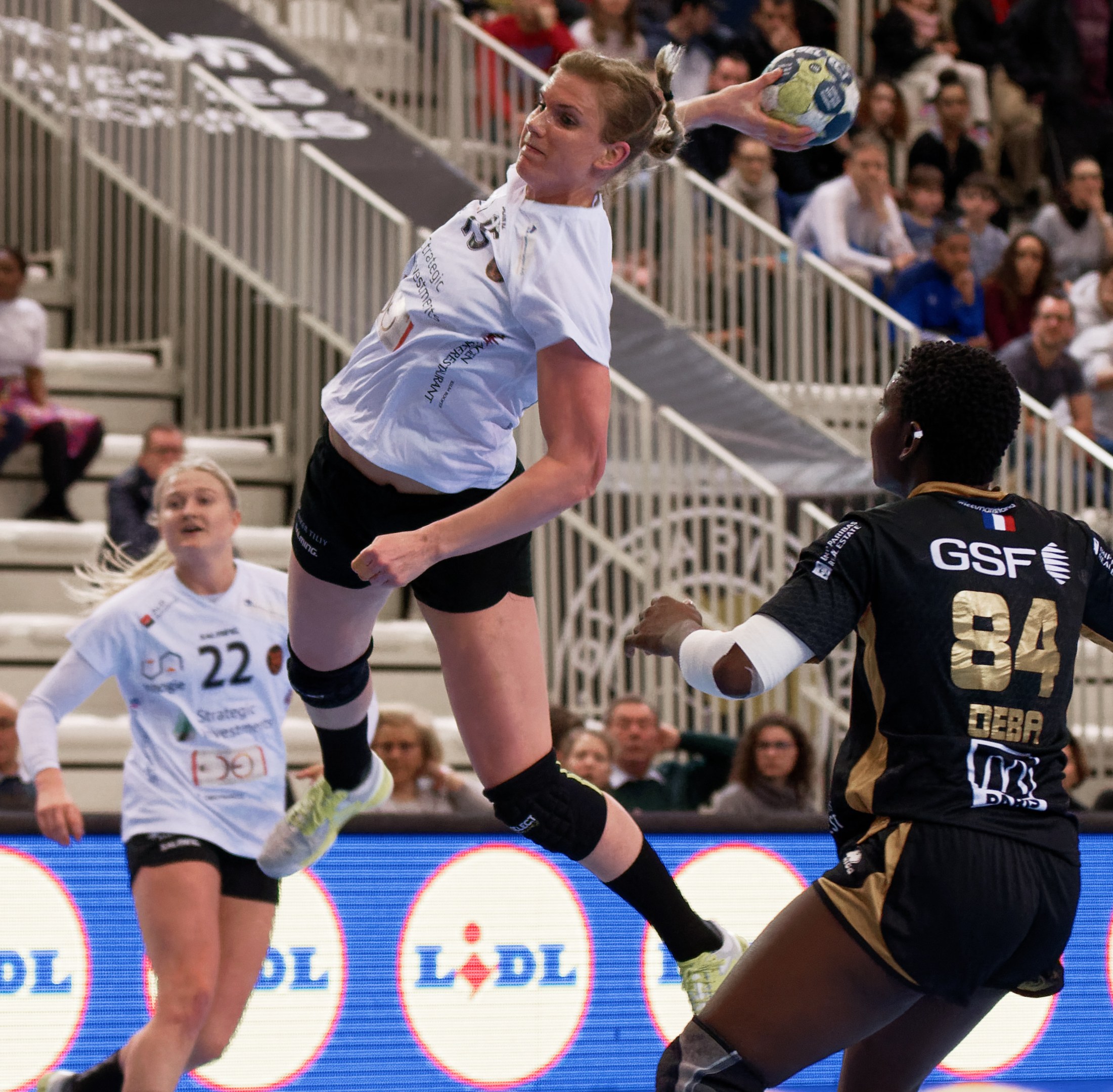 Gruppe von Frauen, die Handball auf einem Platz spielen, mit einem Ball in der Luft, Zuschauern auf Treppen und Geländern und einem "Handball-Weltmeisterschaft 2019 - Tag 2"-Schild im Hintergrund.
