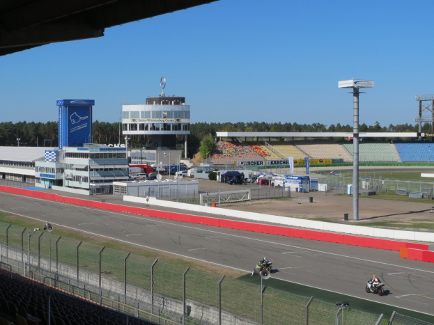 Gruppe von Motorradfahrern auf einer Rennstrecke, umgeben von einem Metallzaun, Gras, Fahrzeugen, einem Gebäude, einem Schild, Pfosten, einem Stadion, einem Dach mit Deckenbeleuchtung, Bäumen und einem bewölkten Himmel.