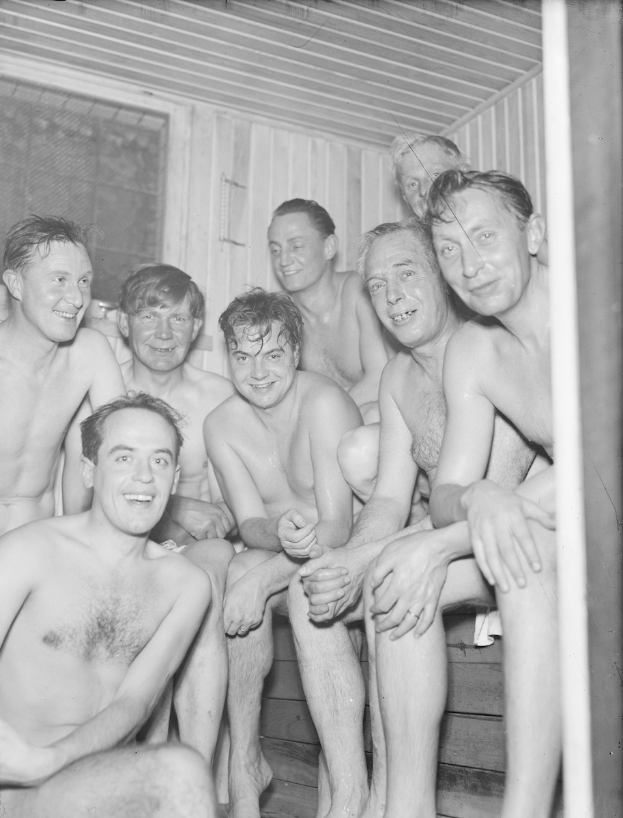 Eine Gruppe von Männern sitzt auf einer Holzbank in einer Sauna, mit einer Wand und einem Fenster im Hintergrund, dargestellt in Schwarz-Weiß.