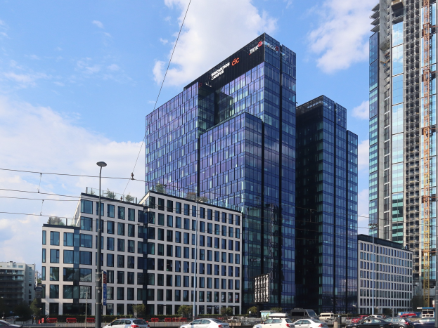 Außenansicht des Deutschen Bank-Hauptsitzes in Frankfurt, Deutschland, mit einem großen Gebäude mit zahlreichen Fenstern, umgeben von städtischer Infrastruktur wie Straßenlaternen, Fahrzeugen, Bäumen und einem bewölkten Himmel.
