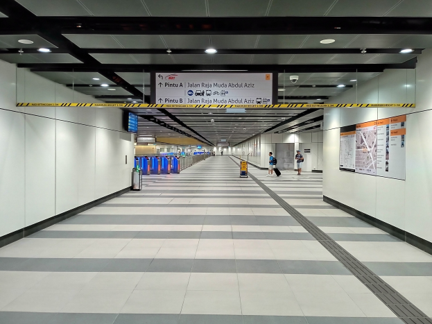 Ein langer Flughafenkorridor mit Menschen, die gehen, Plakate an der rechten Wand, Deckenleuchten, eine Überwachungskamera und ein Gepäckausgabe-Bereich in der Mitte.