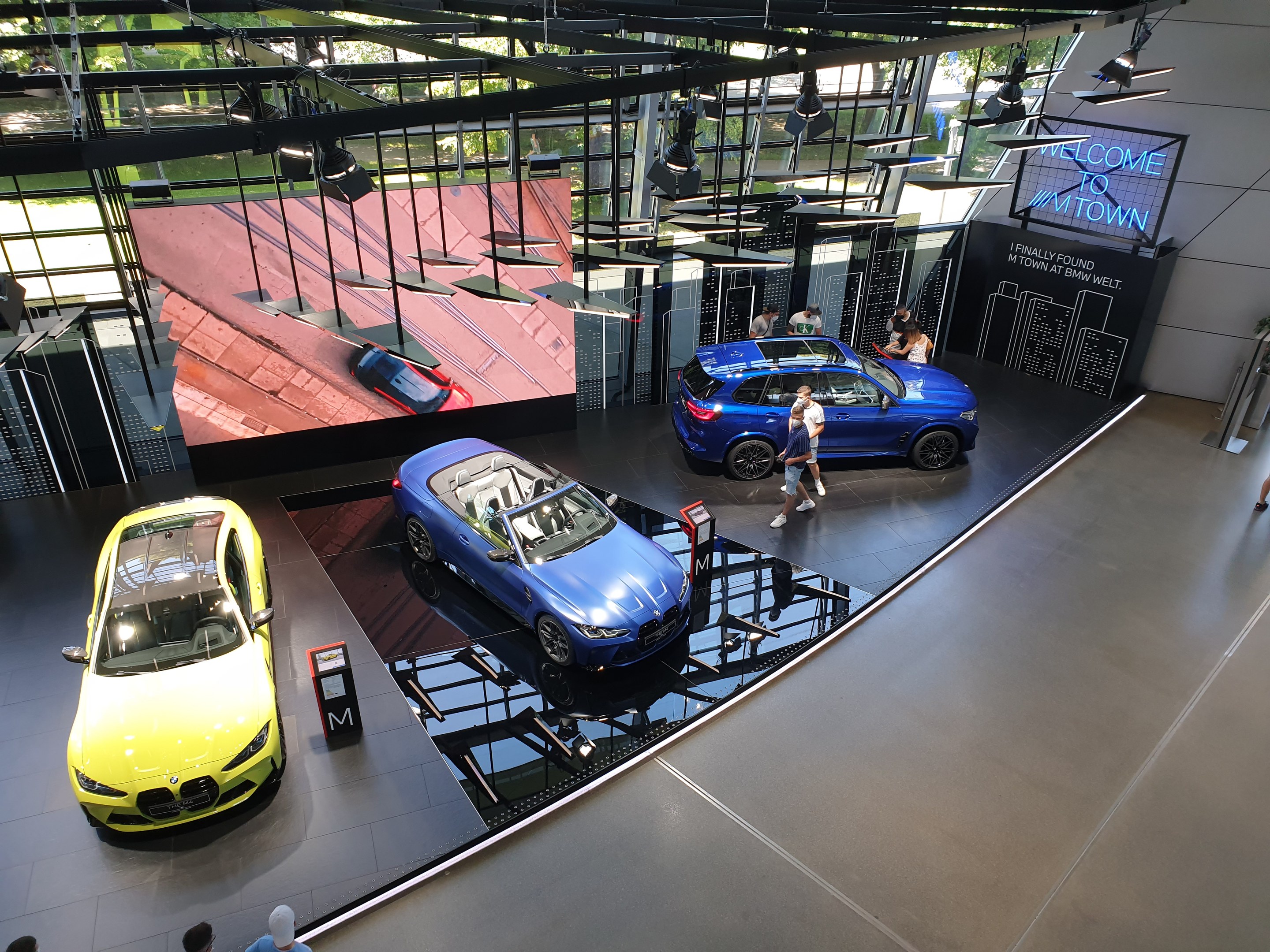 Eine Gruppe von Menschen, die im BMW Museum in München stehen, mit Autos auf der linken Seite und einem Schild auf der rechten Seite, das "BMW Museum München" lautet, mit Blick auf eine städtische Außenansicht durch Glasfenster.