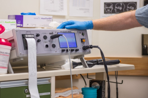 Eine Person in blauen Handschuhen arbeitet an einem medizinischen Gerät an einem unordentlichen Tisch in einem Raum mit Papieren an der Wand, was auf eine Krankenhausumgebung hinweist.