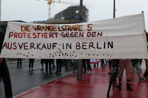 Eine Gruppe von Menschen steht auf einer Straße und hölt ein Transparent mit der Aufschrift "Die Wrangellstraße 5 protestiert gegen den Ausverkauf in Berlin". Im Hintergrund sind Fahrráder, Stäbe, Gebáude und ein Kran zu sehen, öber ihnen der Himmel.