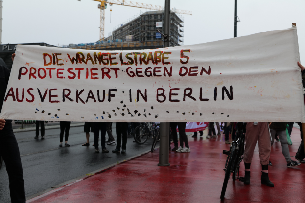 Eine Gruppe von Menschen steht auf einer Straße und hölt ein Transparent mit der Aufschrift "Die Wrangellstraße 5 protestiert gegen den Ausverkauf in Berlin". Im Hintergrund sind Fahrráder, Stäbe, Gebáude und ein Kran zu sehen, öber ihnen der Himmel.