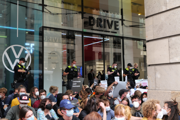 Eine große Gruppe von Menschen mit Masken hält Schilder vor einem Glasgebäude, wobei einige Mützen tragen und Kameras halten.