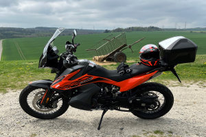 Ein KTM 1290 Super Adventure R Motorrad auf einem Schotterweg geparkt mit einem Helm und einem Gepäckwagen daneben, umgeben von grünem Gras, hohen Bäumen und Strommasten unter einem bewölkten Himmel.