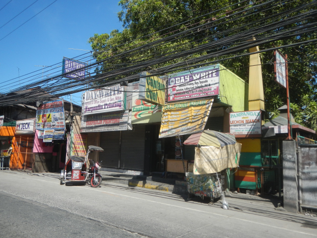 Eine Straßenszene mit einem geparkten Motorrad, Gebäuden, Geschäften, Namenschildern, Strommasten, Kabeln, Bäumen und einem klaren blauen Himmel.