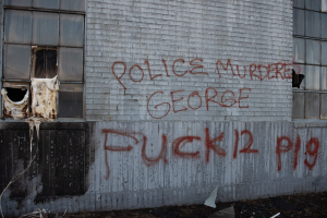 Ein Gebäude mit Graffiti, das "Polizei Mord George Fuck 12 P19" lautet, auf der Seite, mit Fenstern und verschiedenen Gegenständen vorne.