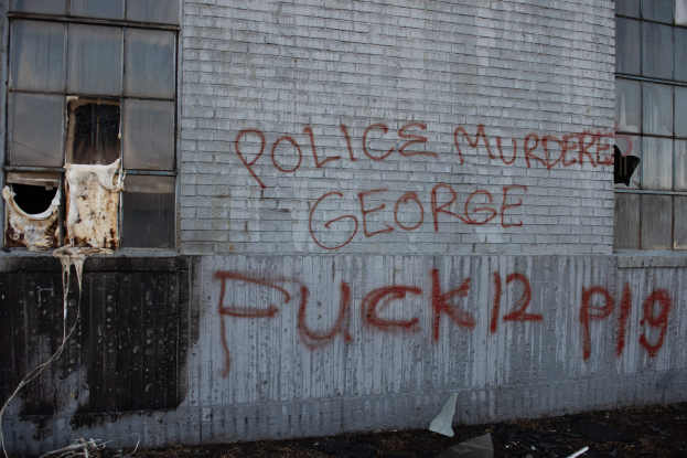 Ein Gebäude mit Graffiti, das "Polizei Mord George Fuck 12 P19" lautet, auf der Seite, mit Fenstern und verschiedenen Gegenständen vorne.