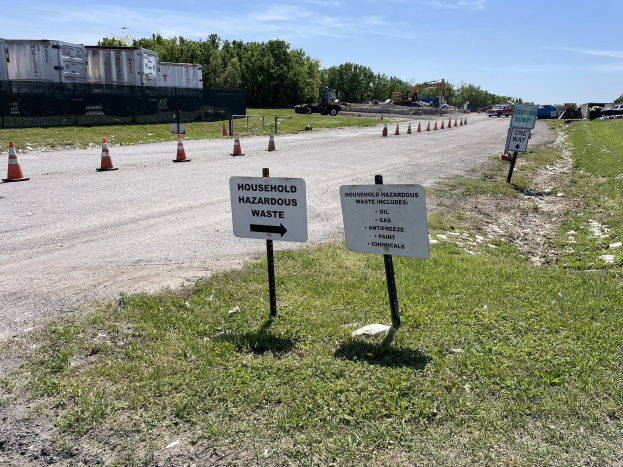 Straßenseite mit Sammelstelle für "Gefährliche Haushaltsabfälle" markiert, mit Gras, Verkehrskegeln, Fahrzeugen, Bäumen und einem bewölkten Himmel im Hintergrund.