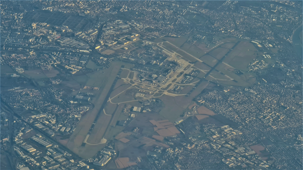 Luftaufnahme eines Flughafens umgeben von städtischer Infrastruktur, einschließlich Gebäuden und Straßen, mit Text unten.