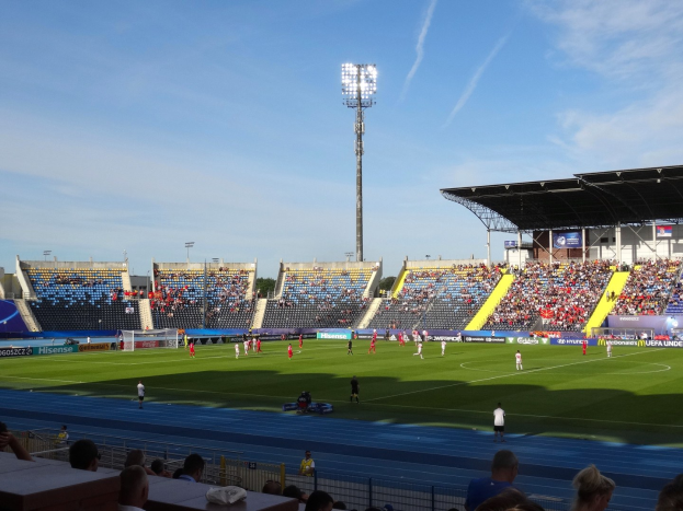 Großes Stadion mit Flutlichtfußballspiel, Zuschauer auf Tribüne, Spieler auf Feld, sichtbarer Himmel.