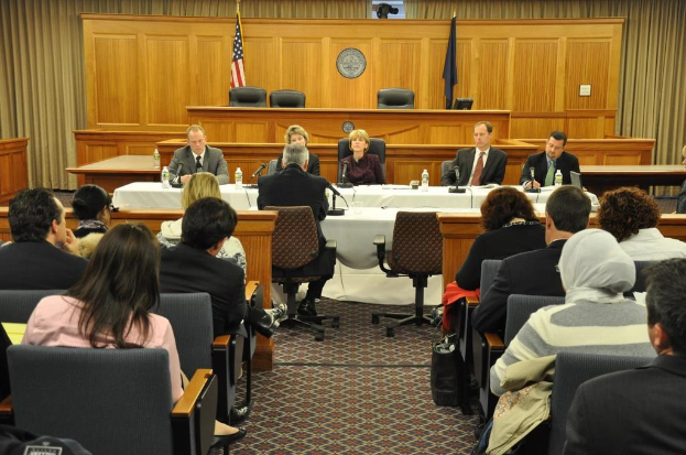Ein Versammlungsraum mit Menschen auf Stühlen, zwei ausländische Flaggen, ein oranger Schreibtisch, Wasserflaschen und ein Mikrofon.