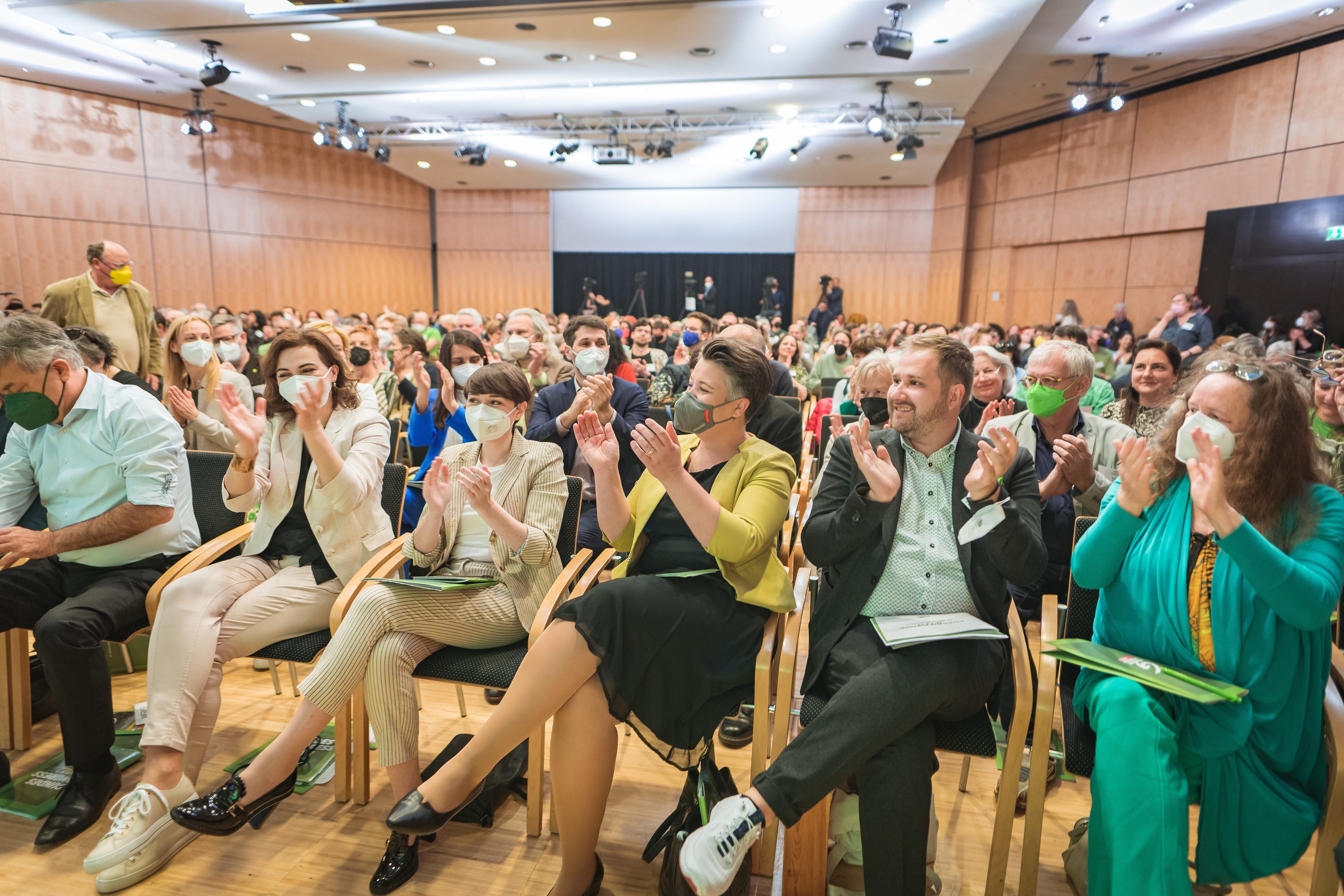 Eine Gruppe von Menschen sitzt in Stühlen vor einem Publikum, einige tragen Masken, mit Taschen auf dem Boden, in einem Konferenzsetting mit einem Bildschirm und Deckenleuchten.