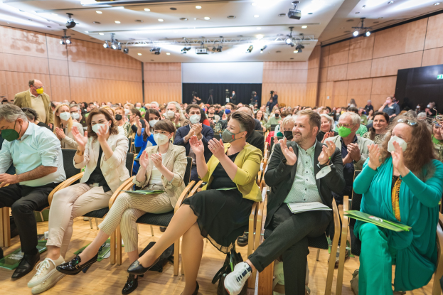 Eine Gruppe von Menschen sitzt in Stühlen vor einem Publikum, einige tragen Masken, mit Taschen auf dem Boden, in einem Konferenzsetting mit einem Bildschirm und Deckenleuchten.
