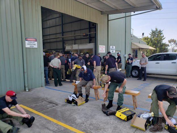 Gruppe von Menschen in militärischer Uniform vor einem Gebäude mit verstreuten Kartonagen, Schuhen und Ausrüstung, einige sitzen auf Bänken, andere stehen, mit Fahrzeugen, Bäumen und Gebäuden im Hintergrund unter einem klaren blauen Himmel.