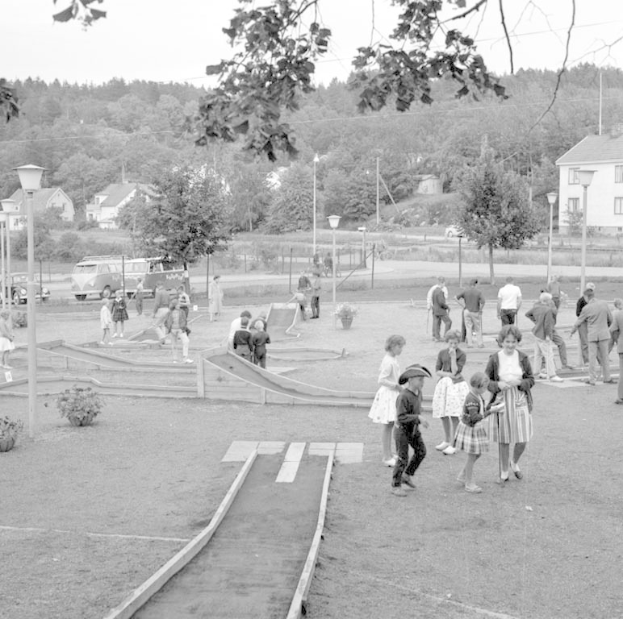 Schwarz-weißes Bild von Menschen, die Minigolf in einem Park mit üppiger Vegetation, Laternen, geparkten Fahrzeugen und Gebäuden im Hintergrund spielen.