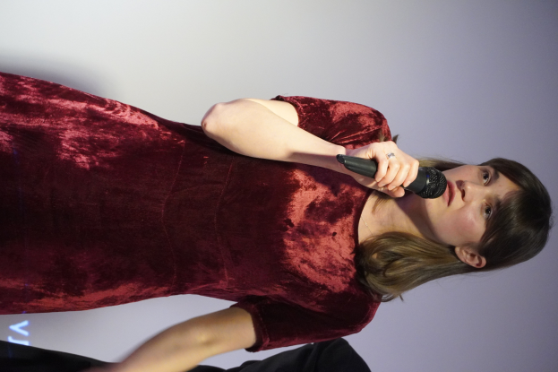 Eine Frau in einem roten Kleid steht selbstbewusst auf einer Bühne, hält ein Mikrofon und hat eine stylische Bob-Frisur und natürliches Make-up.