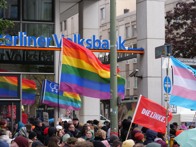 Eine große Gruppe von Menschen mit Fahnen und Schildern steht vor einem Gebäude, mit einem Mast im Vordergrund und Bäumen auf beiden Seiten, bei einer Christopher Street Parade in Berlin.
